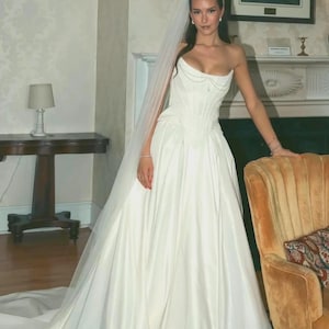 Peut inclure: Une robe de mariée blanche longue jusqu'au sol avec un corsage bustier à baleines verticales et un détail drapé au décolleté. La robe présente une jupe ample avec une traîne, accompagnée d'un long voile blanc transparent.