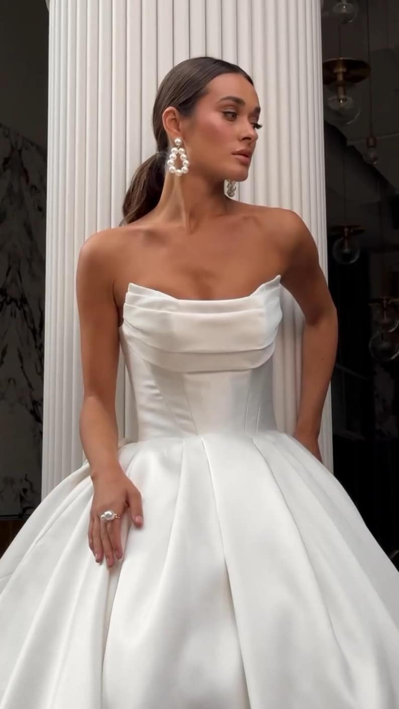 May include: A strapless white satin wedding dress with a fitted bodice and a full skirt. The dress has a simple, elegant design and is perfect for a formal wedding.