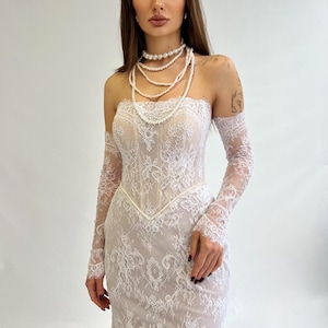 Könnte beinhalten: Ein trägerloses weißes Spitzenkleid mit langen, durchsichtigen Spitzenärmeln. Das Kleid hat ein eng anliegendes Oberteil und einen fließenden Rock. Eine mehrreihige Perlenkette schmückt den Hals. Der Stil ist elegant, für eine Hochzeit oder einen besonderen Anlass.