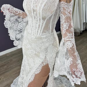May include: White lace wedding dress with a corset bodice and off-the-shoulder sleeves. The dress features a high slit and a flowing skirt, showcasing intricate floral lace detailing. The sleeves have a flared design.