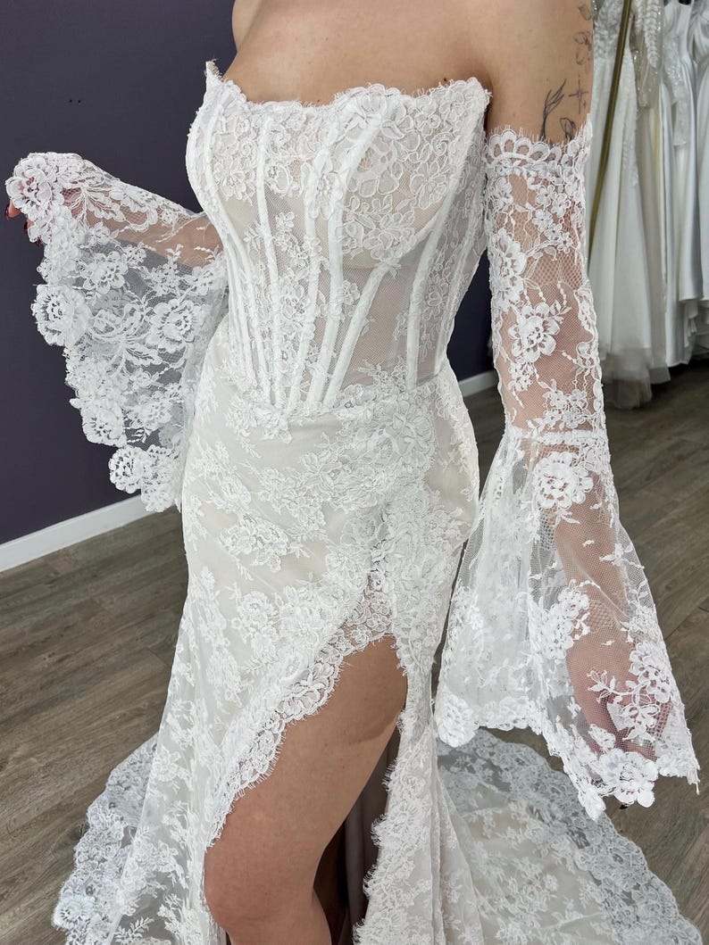 May include: White lace wedding dress with a corset bodice and off-the-shoulder sleeves. The dress features a high slit and a flowing skirt, showcasing intricate floral lace detailing. The sleeves have a flared design.