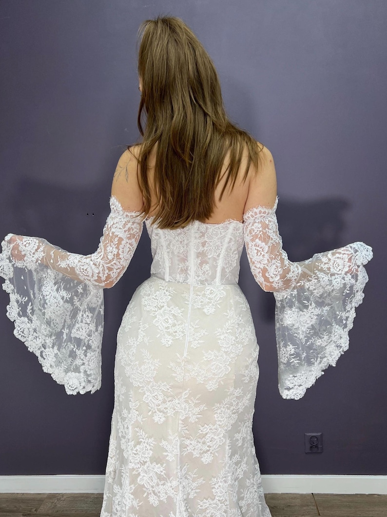 May include: A white lace wedding dress with off-the-shoulder sleeves and a fitted bodice. The dress features intricate floral lace detailing throughout, with flared sleeves. The back of the dress is visible, showcasing the design and fit.