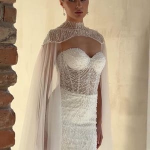 May include: A white wedding dress with a beaded bodice and a sheer cape. The dress features a sweetheart neckline and intricate beadwork. The cape is made of a delicate, flowing fabric. The dress is elegant and suitable for a wedding.