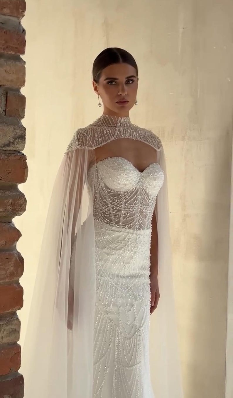 May include: A white wedding dress with a beaded bodice and a sheer cape. The dress features a sweetheart neckline and intricate beadwork. The cape is made of a delicate, flowing fabric. The dress is elegant and suitable for a wedding.