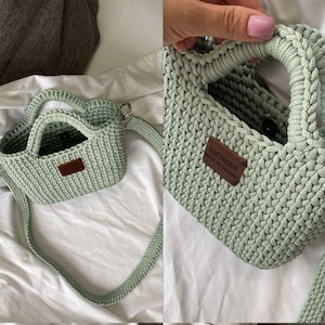 Peut inclure: Un sac à main vert clair, crocheté à la main, de forme rectangulaire, avec une poignée supérieure et une longue bandoulière réglable. Une petite étiquette en cuir marron avec le texte "NIETKOVA" est attachée à l'avant. Le sac est fait d'un fil épais et texturé.