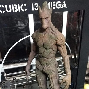 Könnte beinhalten: Eine 3D-gedruckte Figur von Groot aus Guardians of the Galaxy. Die Figur steht auf einem Holzsockel und ist in Braun- und Grüntönen bemalt. Die Figur steht vor einem 3D-Drucker mit dem Text "CUBIC 13 MEGA" auf dem Drucker.
