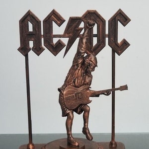 Puede incluir: Estatua de color bronce de un miembro de la banda AC/DC tocando la guitarra, con el logotipo de la banda encima. La figura está sobre una base circular con la inscripción "STIFF UPPER LIP". La estatua mide aproximadamente 20 cm de altura.