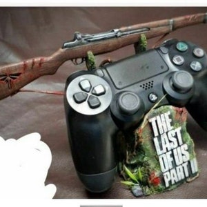 Peut inclure: Une manette de jeu PlayStation 4 noire avec un fusil en bois posé dessus. La manette est posée sur un socle avec les mots "The Last of Us Part II" imprimés dessus.