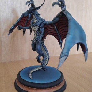 May include: A blue and gray dragon figurine with red wings, standing on a round base with a blue and black design. The dragon has a long tail and sharp claws.