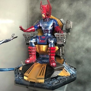 Puede incluir: Una figura de acción detallada del personaje de cómics de Marvel, Galactus, sentado en un trono con una base azul y dorada. La figura lleva un traje rojo y azul con un casco plateado y sostiene un rayo de energía azul en su mano derecha.
