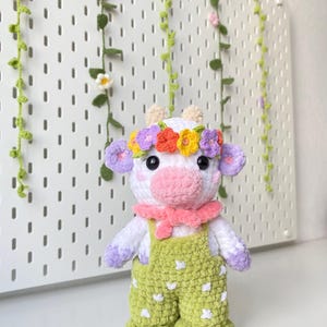 May include: A white crocheted cow plush toy wearing a flower crown, a pink scarf, and green overalls with white polka dots. The toy is standing in front of a white pegboard with green vines and flowers.