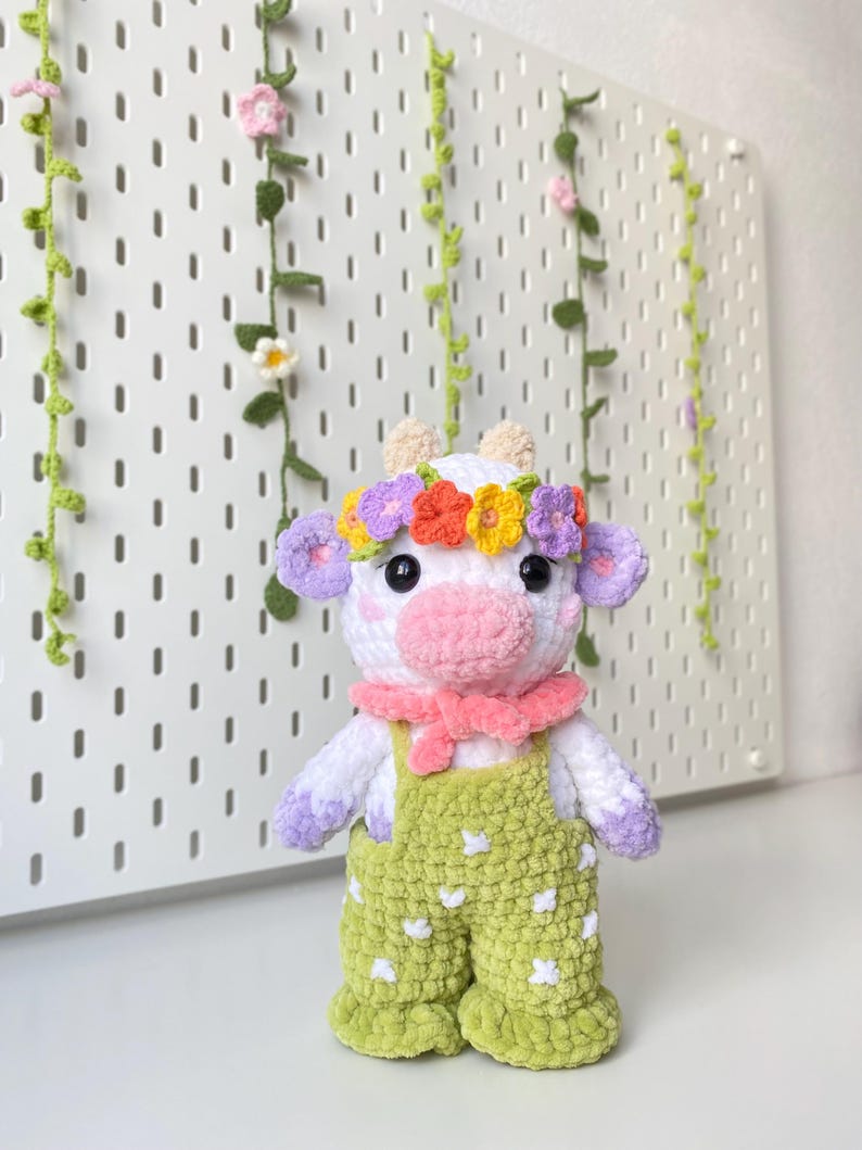 May include: A white crocheted cow plush toy wearing a flower crown, a pink scarf, and green overalls with white polka dots. The toy is standing in front of a white pegboard with green vines and flowers.