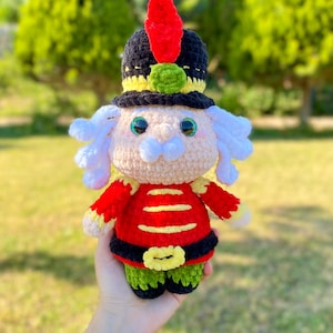 May include: Crocheted nutcracker doll in red, black, and green. The doll has a black hat with a red feather and a green ball. The doll has white hair and a white mustache. The doll is wearing a red jacket with yellow stripes and a black belt.