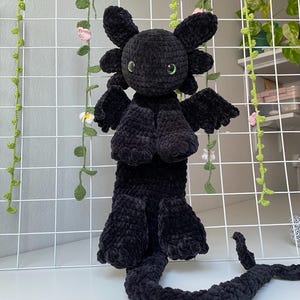 Puede incluir: Juguete de peluche de dragón negro de crochet con ojos verdes. El dragón tiene una cola larga y está de pie sobre una superficie blanca.