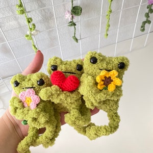 May include: Three green crocheted frog plushies holding different flowers. The frog on the left is holding a pink flower, the middle frog is holding a red heart, and the frog on the right is holding a yellow sunflower.