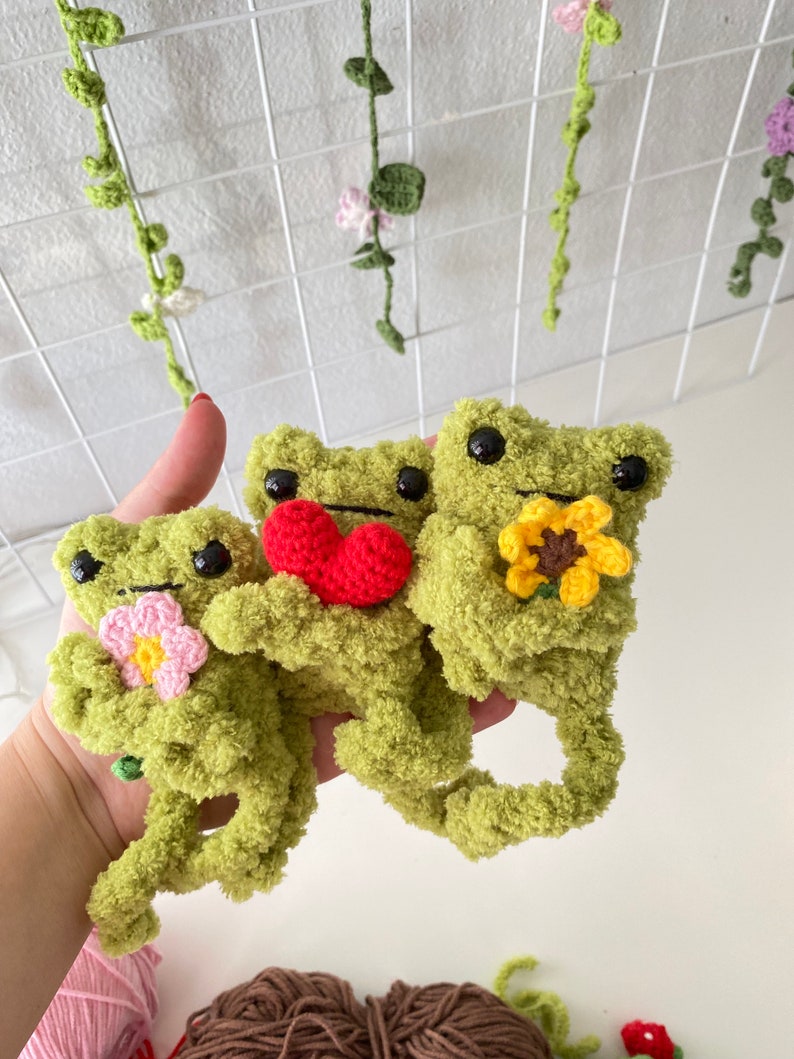 May include: Three green crocheted frog plushies holding different flowers. The frog on the left is holding a pink flower, the middle frog is holding a red heart, and the frog on the right is holding a yellow sunflower.