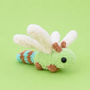 May include: A crocheted dragonfly plush toy featuring a light green head and body, four white wings, and brown accents on the head and tail. The tail has alternating bands of light blue and brown, and the toy has two small black plastic eyes.