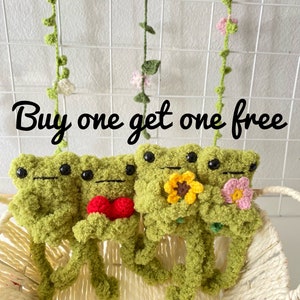 May include: Buy one get one free. Three crocheted green frog plushies with different accessories. The first frog is holding a red heart, the second frog is holding a yellow sunflower, and the third frog is holding a pink flower.