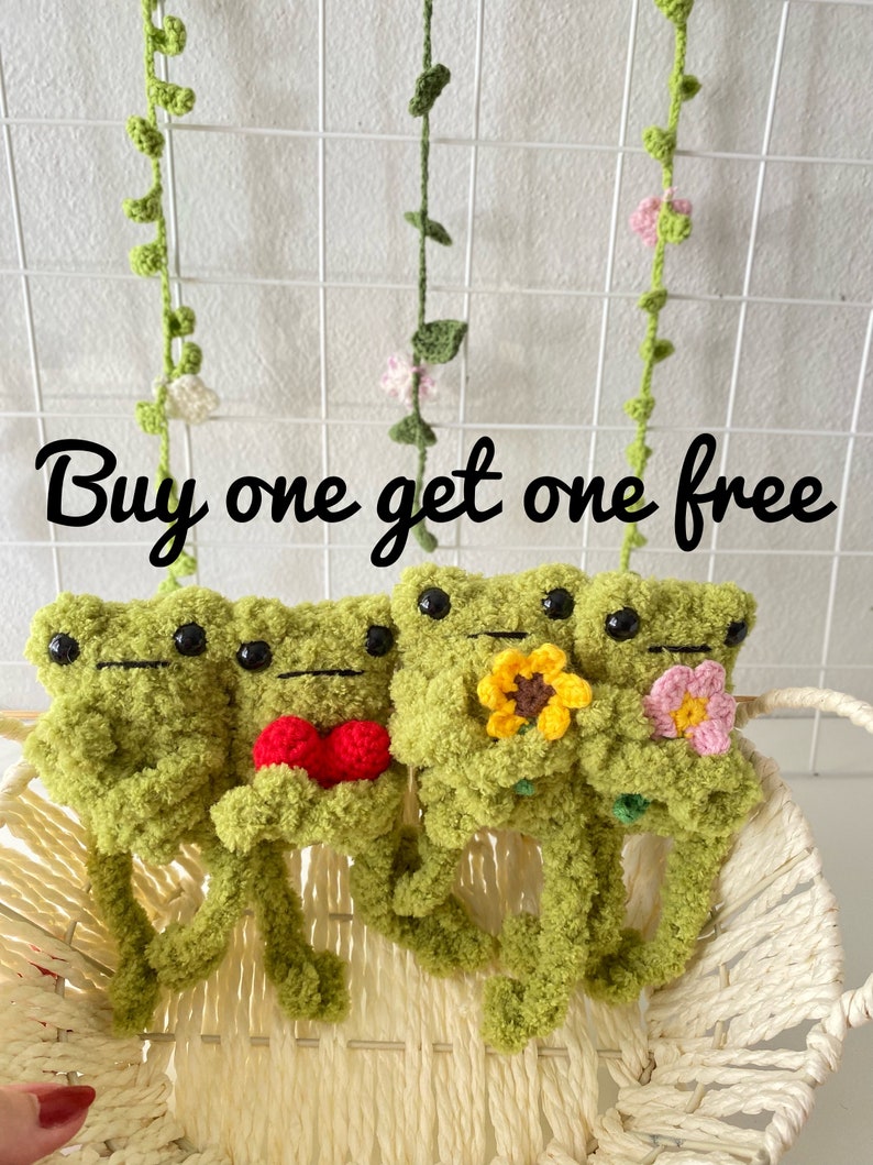 May include: Buy one get one free. Three crocheted green frog plushies with different accessories. The first frog is holding a red heart, the second frog is holding a yellow sunflower, and the third frog is holding a pink flower.