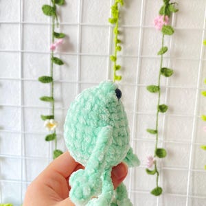May include: A crocheted green frog plush toy with black eyes. The frog is sitting on a hand.