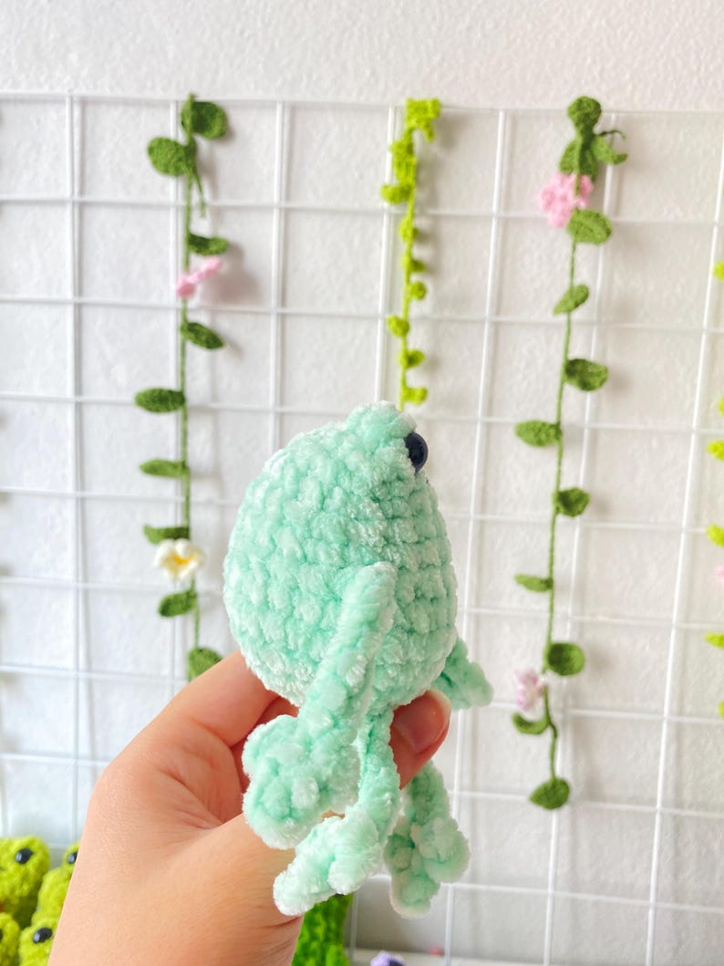 May include: A crocheted green frog plush toy with black eyes. The frog is sitting on a hand.
