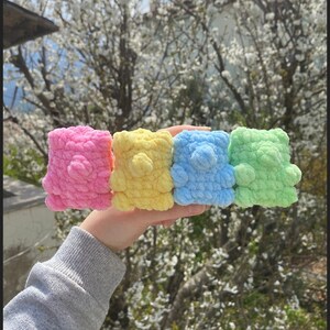 May include: Four crocheted teddy bears in pink, yellow, blue, and green. The bears are lined up in a row and are being held by a hand.