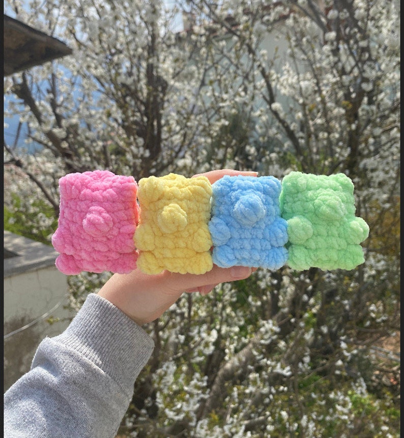 May include: Four crocheted teddy bears in pink, yellow, blue, and green. The bears are lined up in a row and are being held by a hand.