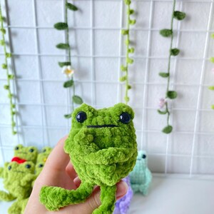 May include: A green crocheted frog plush toy with black eyes. The frog is sitting on a hand with other crocheted frog toys in the background.