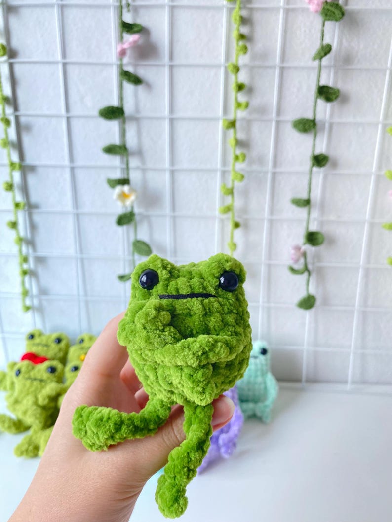 May include: A green crocheted frog plush toy with black eyes. The frog is sitting on a hand with other crocheted frog toys in the background.