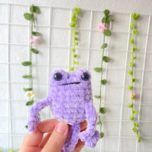 May include: A purple crocheted frog toy with black eyes. The frog is sitting in a person's hand.