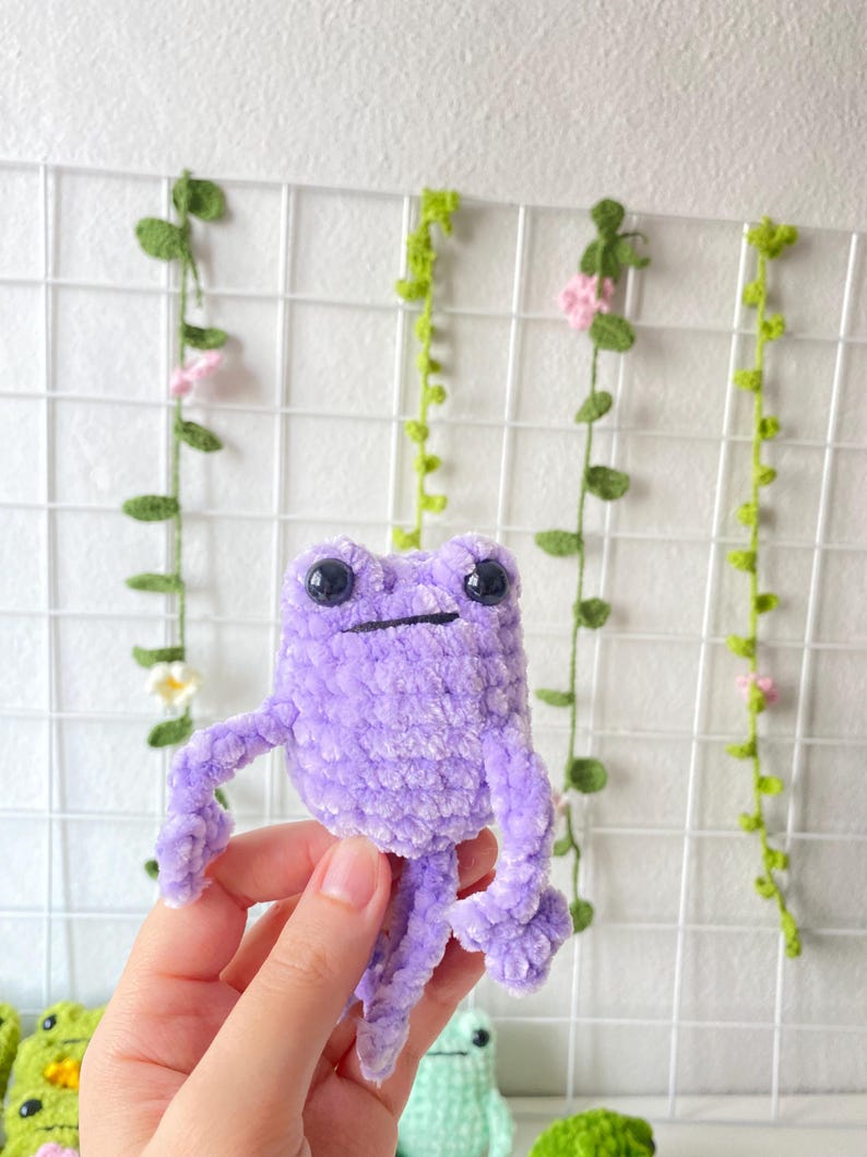 May include: A purple crocheted frog toy with black eyes. The frog is sitting in a person's hand.