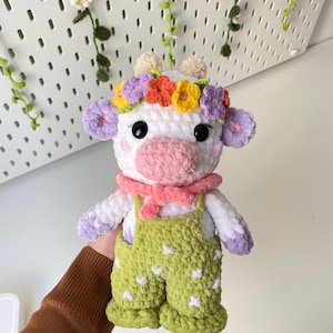 May include: A white crocheted cow plush toy wearing a flower crown and green overalls with white polka dots. The cow has pink ears, a pink nose, and black eyes.
