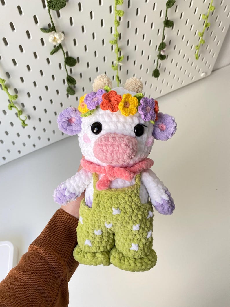 May include: A white crocheted cow plush toy wearing a flower crown and green overalls with white polka dots. The cow has pink ears, a pink nose, and black eyes.