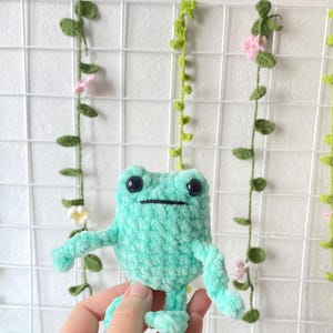 May include: A crocheted green frog plush toy with black eyes and a black mouth. The frog is sitting on a hand.