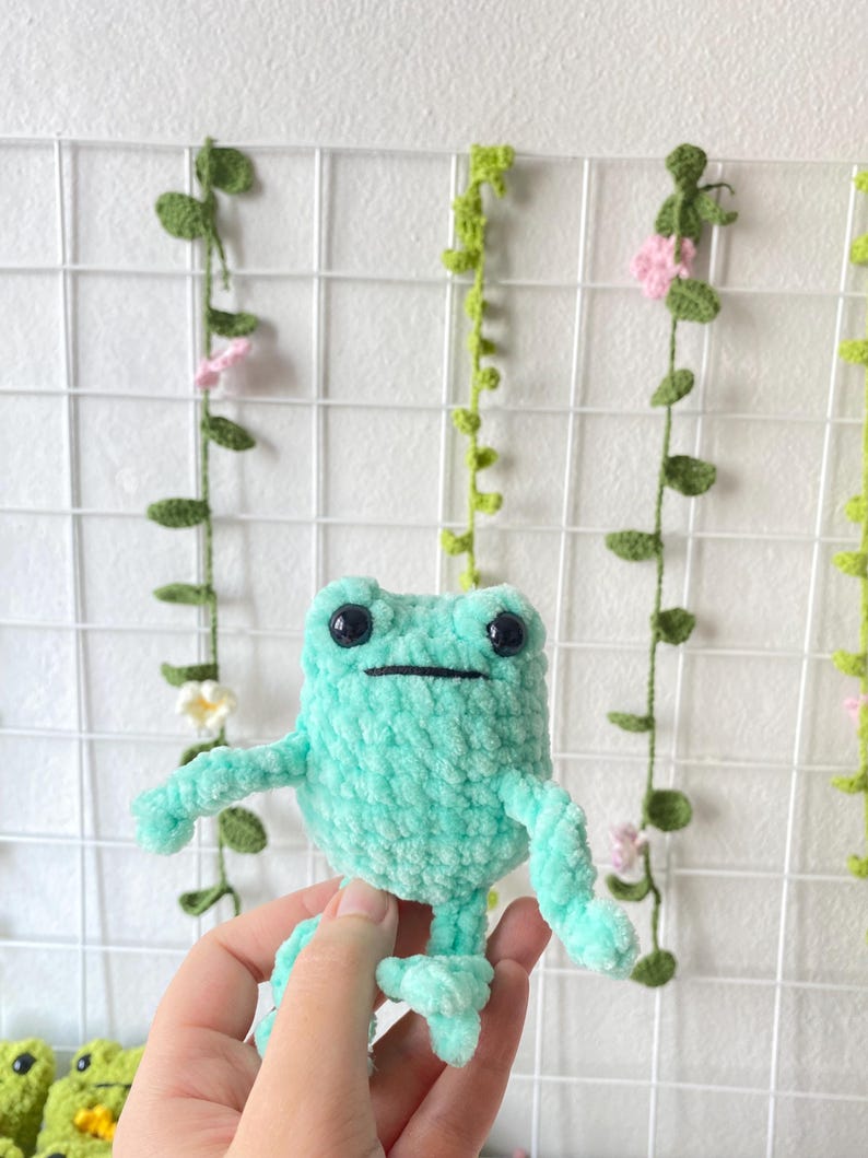 May include: A crocheted green frog plush toy with black eyes and a black mouth. The frog is sitting on a hand.