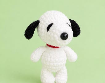 May include: A small, white, crocheted plush toy resembling a beagle. The figure features black, floppy, crocheted ears, a round black plastic nose, and embroidered black eyes. A thin, bright red yarn collar encircles the neck of the figure.
