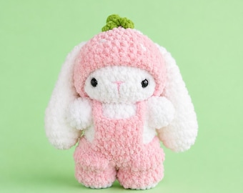 May include: A white crochet bunny plush toy wearing a pink strawberry-patterned hat with a green stem on top and pink crochet overalls. The bunny has long white ears, black bead eyes, and a pink stitched nose, crafted from soft yarn.