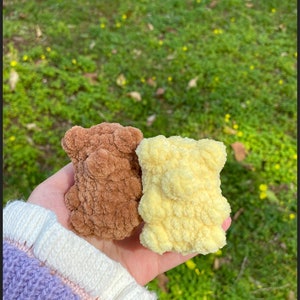May include: Two small crocheted teddy bears, one brown and one yellow, are held in a hand. The bears have a fluffy texture.