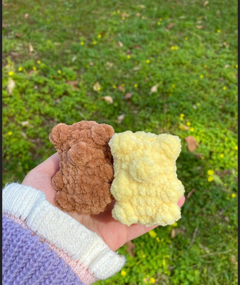 May include: Two small crocheted teddy bears, one brown and one yellow, are held in a hand. The bears have a fluffy texture.