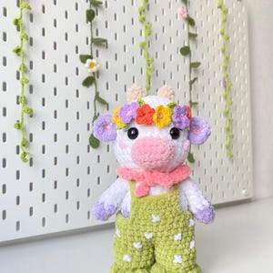May include: A crocheted plush toy cow wearing a flower crown, a pink scarf, and green overalls with white polka dots.