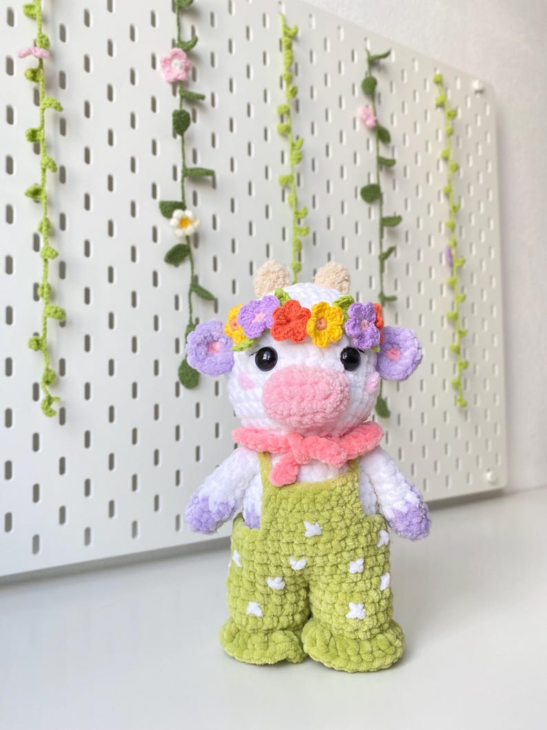 May include: A crocheted plush toy cow wearing a flower crown, a pink scarf, and green overalls with white polka dots.