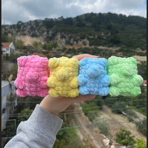 May include: Four crocheted teddy bears in different colors: pink, yellow, blue, and green. The bears are small and fluffy, and they are arranged in a row.