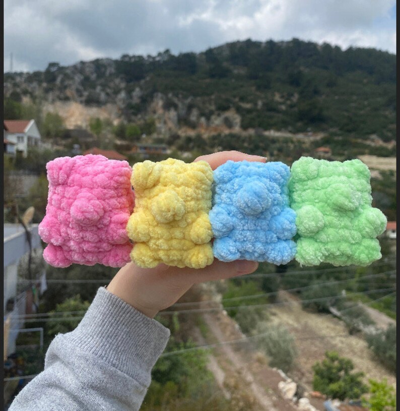May include: Four crocheted teddy bears in different colors: pink, yellow, blue, and green. The bears are small and fluffy, and they are arranged in a row.