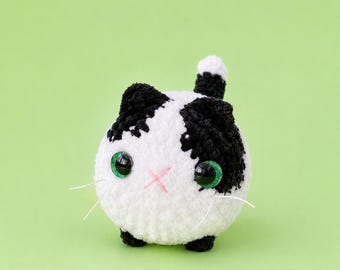 May include: A round, crocheted plush cat with white and black yarn. The cat features large, glossy green eyes, a small pink stitched 'x' for a nose, and thin white whiskers. A small black and white tail extends upward from the top of the body.