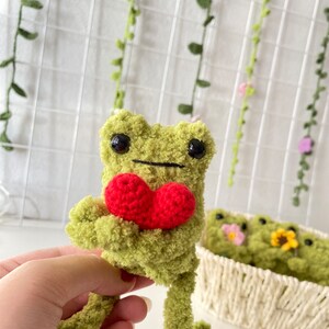 May include: A green crocheted frog holding a red crocheted heart. The frog has black eyes and a black mouth. The frog is sitting on a hand.