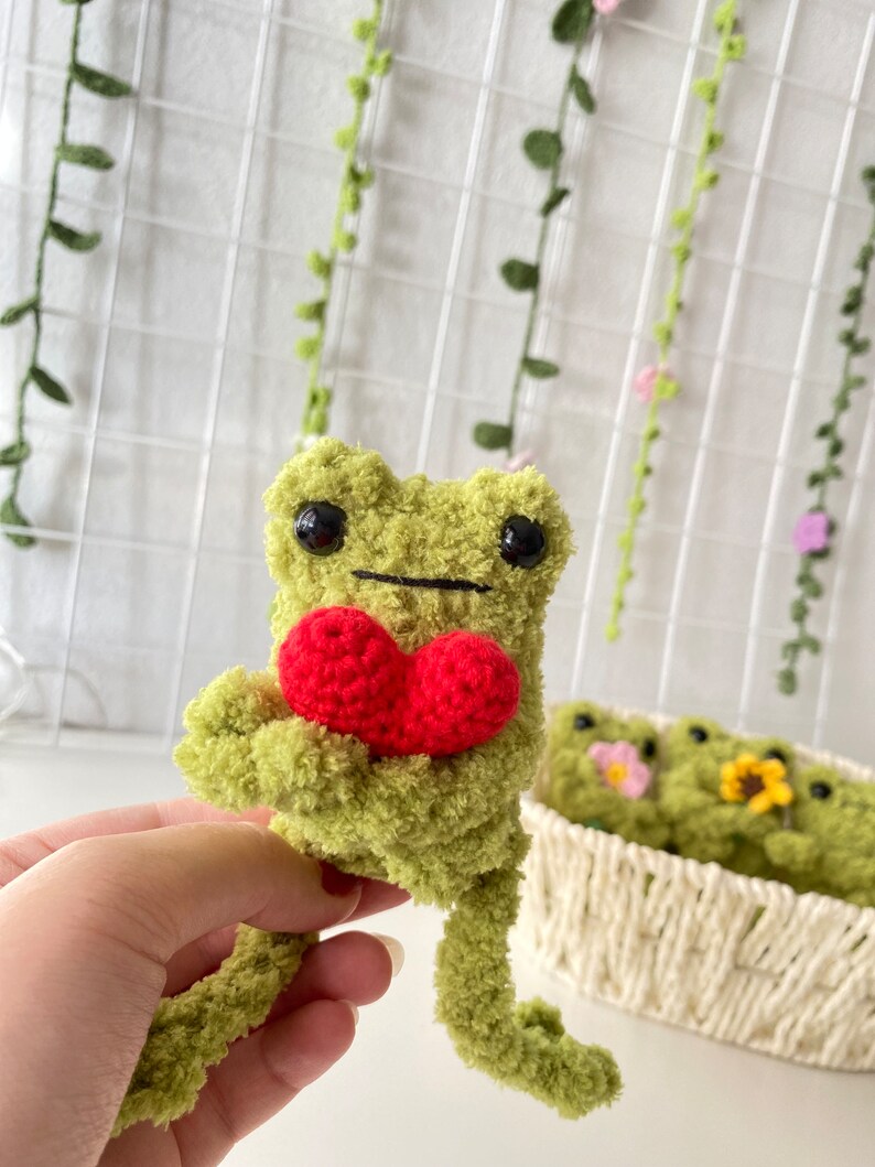 May include: A green crocheted frog holding a red crocheted heart. The frog has black eyes and a black mouth. The frog is sitting on a hand.