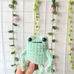 May include: A crocheted green frog plush toy with black eyes. The frog is sitting on a hand.