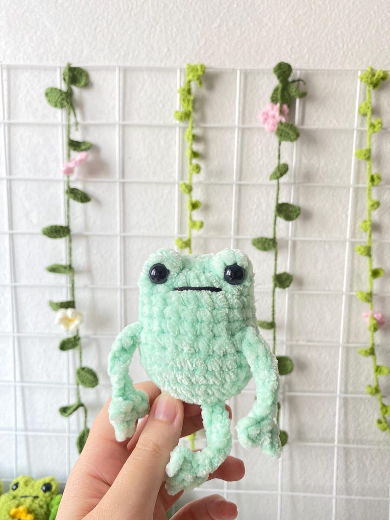 May include: A crocheted green frog plush toy with black eyes. The frog is sitting on a hand.