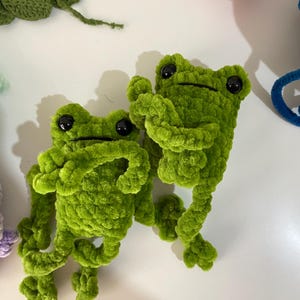 May include: Two green crocheted frog plushies with black eyes. The frogs are sitting on a white surface.