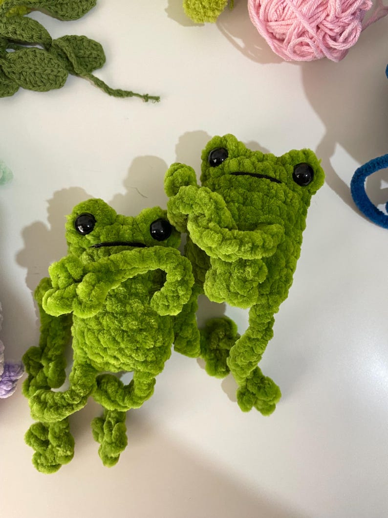 May include: Two green crocheted frog plushies with black eyes. The frogs are sitting on a white surface.